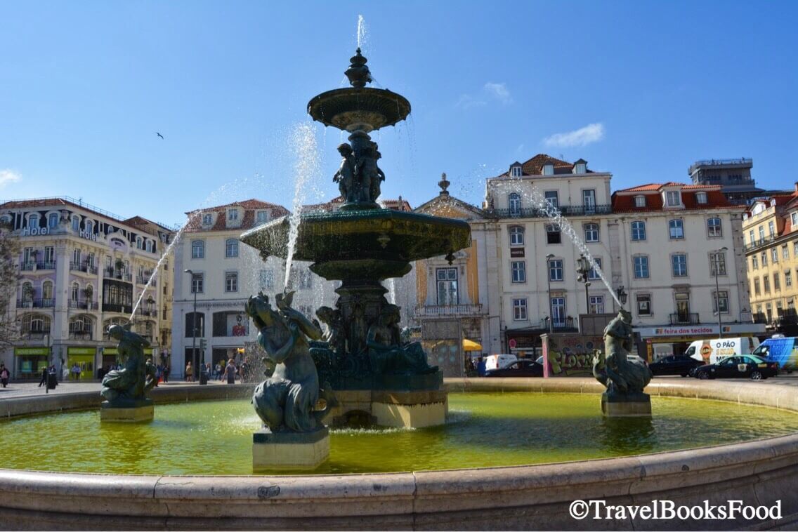A photo of a fountain in Rossio Square in Lisbon surrounded by buildings.