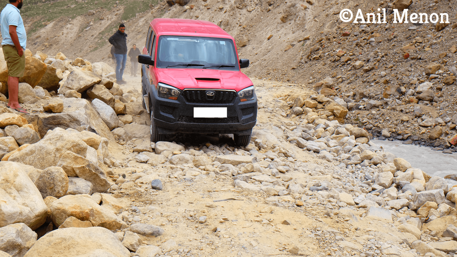 A car trying to navigate the treacherous mountainous gravel roads of the Himalayas with people looking on.
