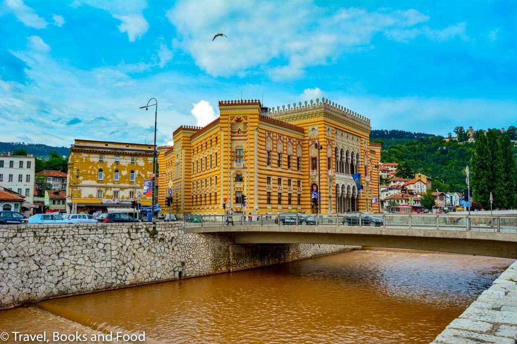 Sarajevo City Hall which is an orange/dark pink building surrounded by a dirty river. One of your stops in your Sarajevo itinerary
