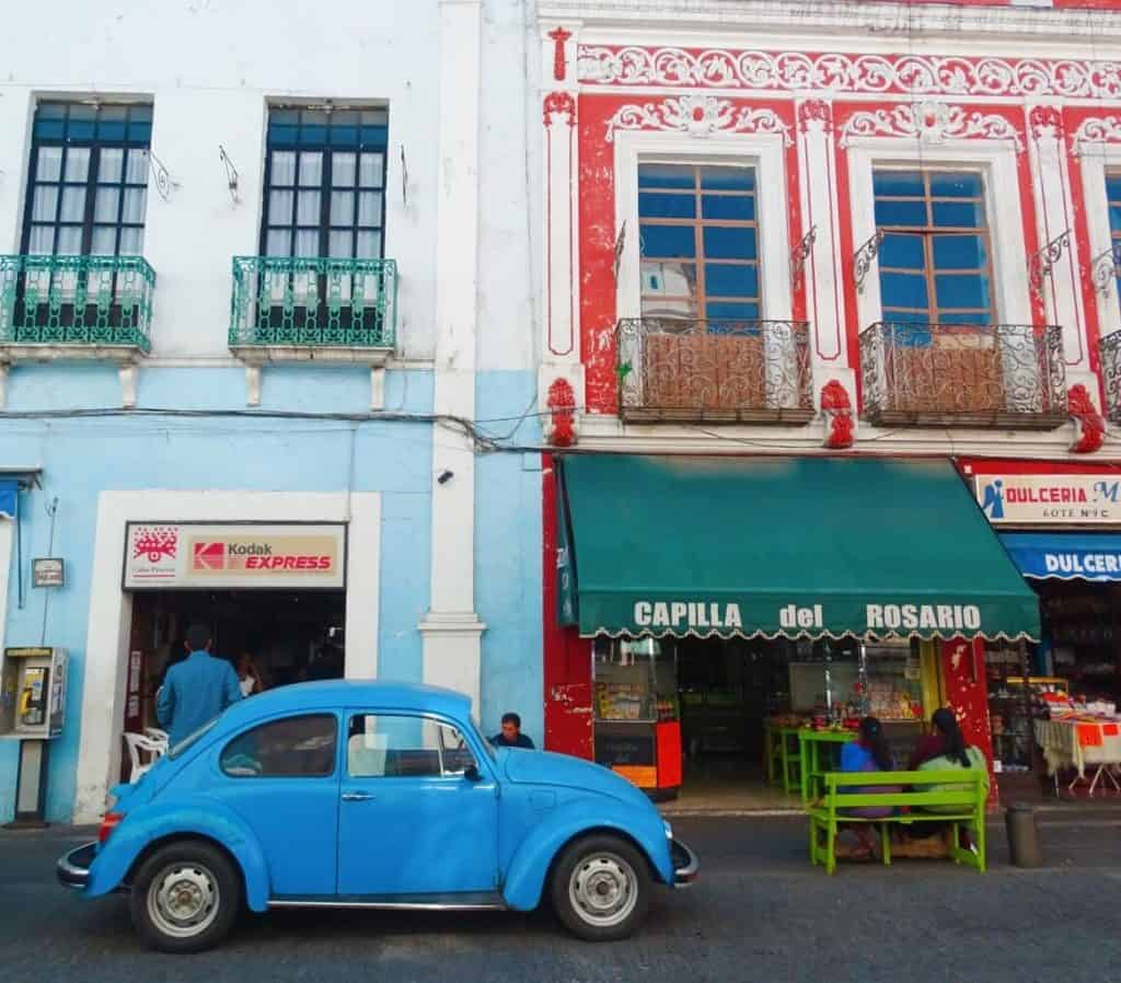 A blue Volkswagon Beetle parked in front of some colorful shops with Baroque architecture in Puebla